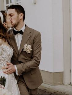 Braut und Bräutigam küssen sich. Im Hintergrund sieht man die Hochzeitslocation. Die Braut trägt ein weißes Spitzenkleid und der Bräutigam einen braunen Anzug.