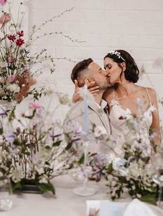 Verliebtes Brautpaar sitzt an Hochzeitstafel, die dekoriert ist mit violetten, weißen und rosa Blumen.