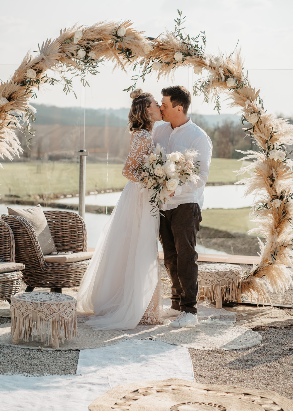 Das Hochzeitspaar, eine Frau mit langem weißen Spitzenkleid und ein Mann mit dunkler Hose und weißem Hemd stehen im Außenbereich der Location und küssen sich unter dem Traubogen bei der Pampasgras Hochzeit.