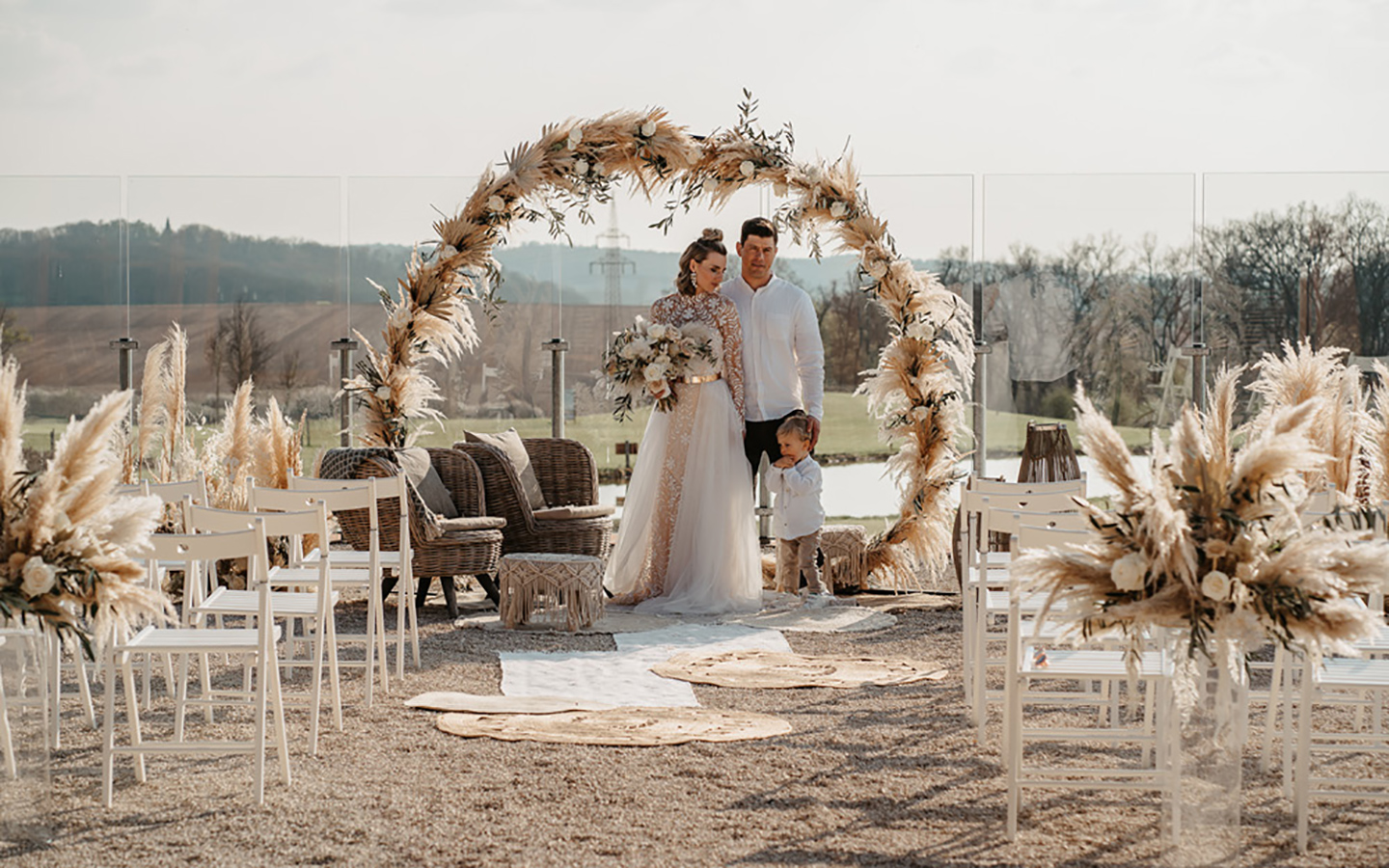 Das Hochzeitspaar steht zusammen mit dem ca. dreijährigen Sohn unter dem Traubogen aus Pampasgras im Außenbereich der Hochzeitslocation. Die weißen Stühle sind so angeordnet, dass die Blicke der Gäste auf das Paar gerichtet sein werden.