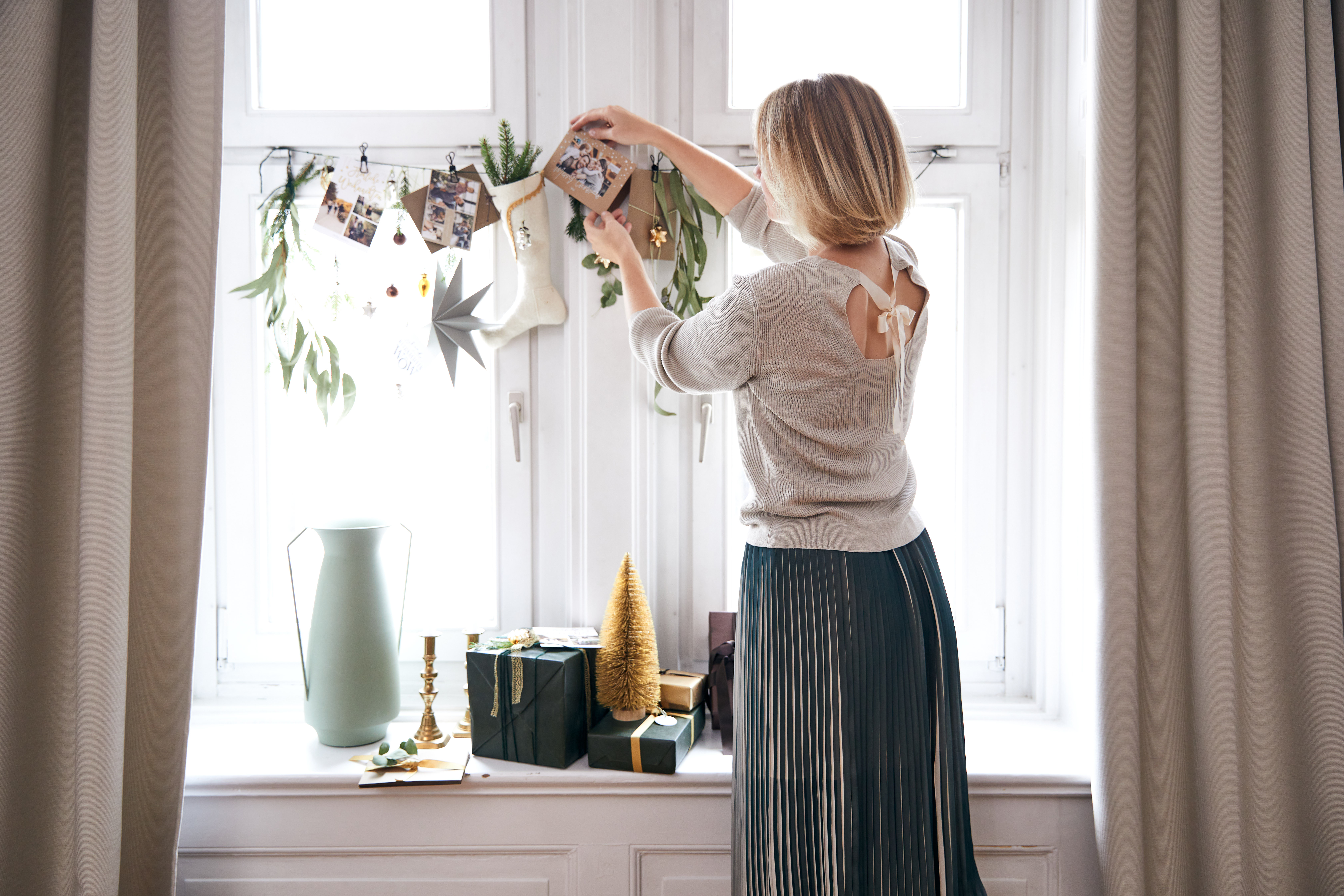 Elegant angezogene Frau schmückt ein Fenster weihnachtlich mit Weihnachtskarten und Blätttergirlande