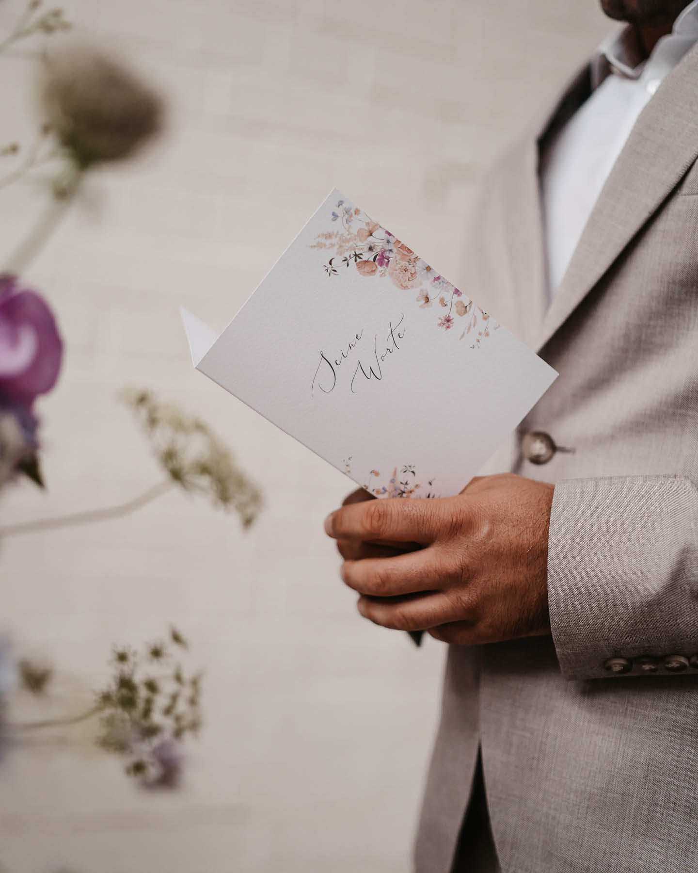 Bräutigam im hellen Anzug hält Ehegelübde als Karte mit floralen Elementen in der Hand.