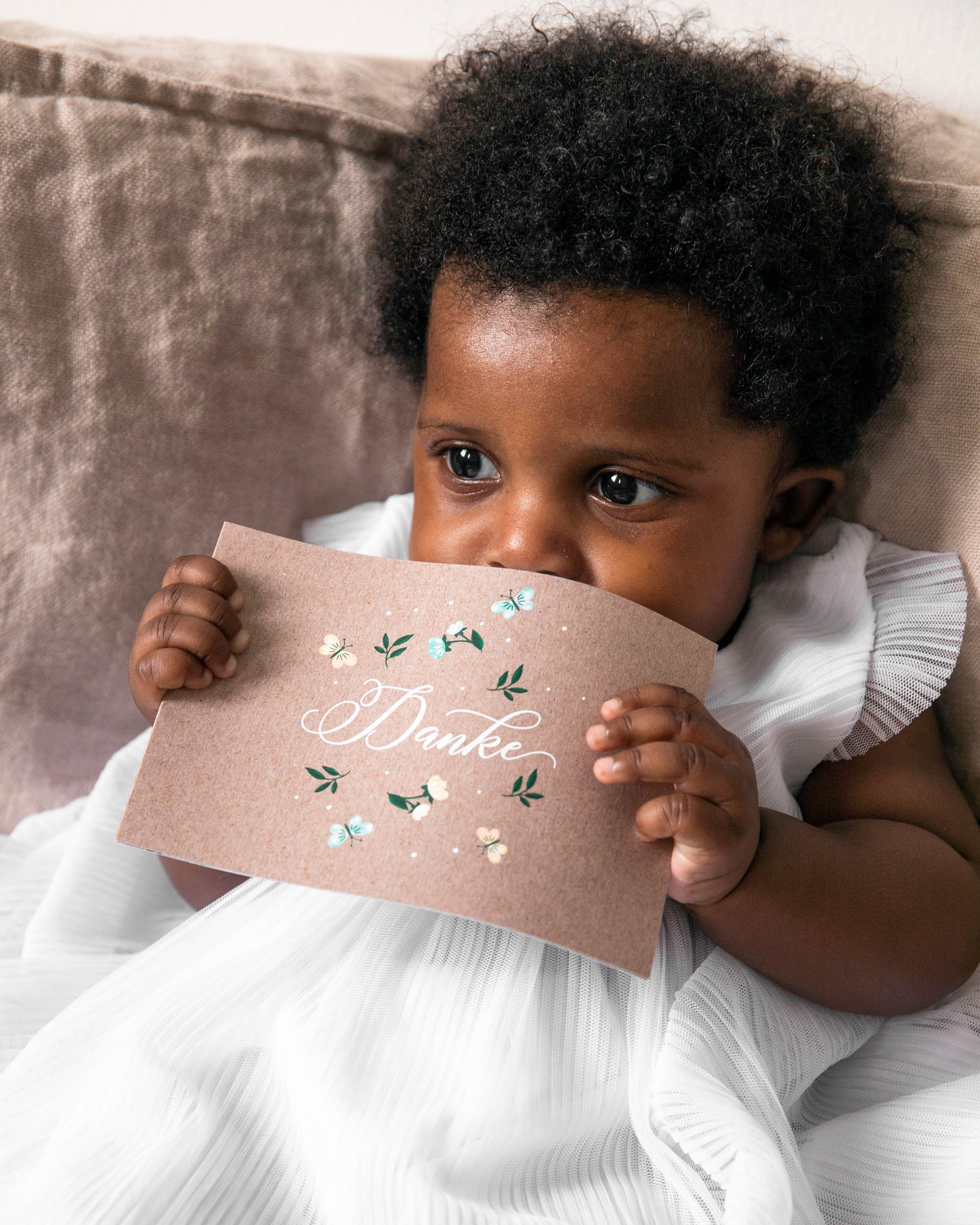 Baby sitzt im Taufkleid auf Sofa und hält in den Hände eine Dankeskarte zur Taufe