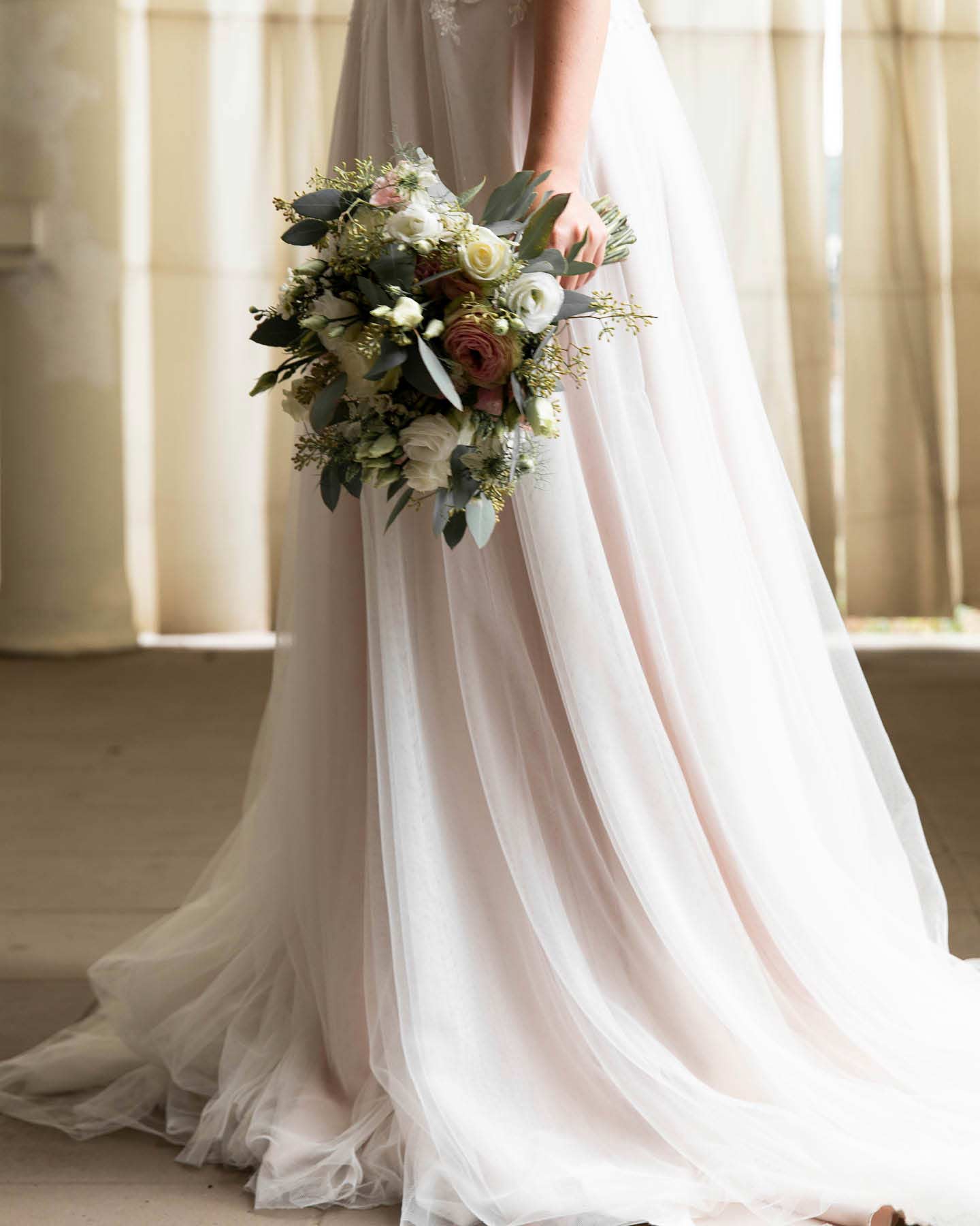Braut hält Brautstrauß vor den fließenden Tüllrock ihres Brautkleides.