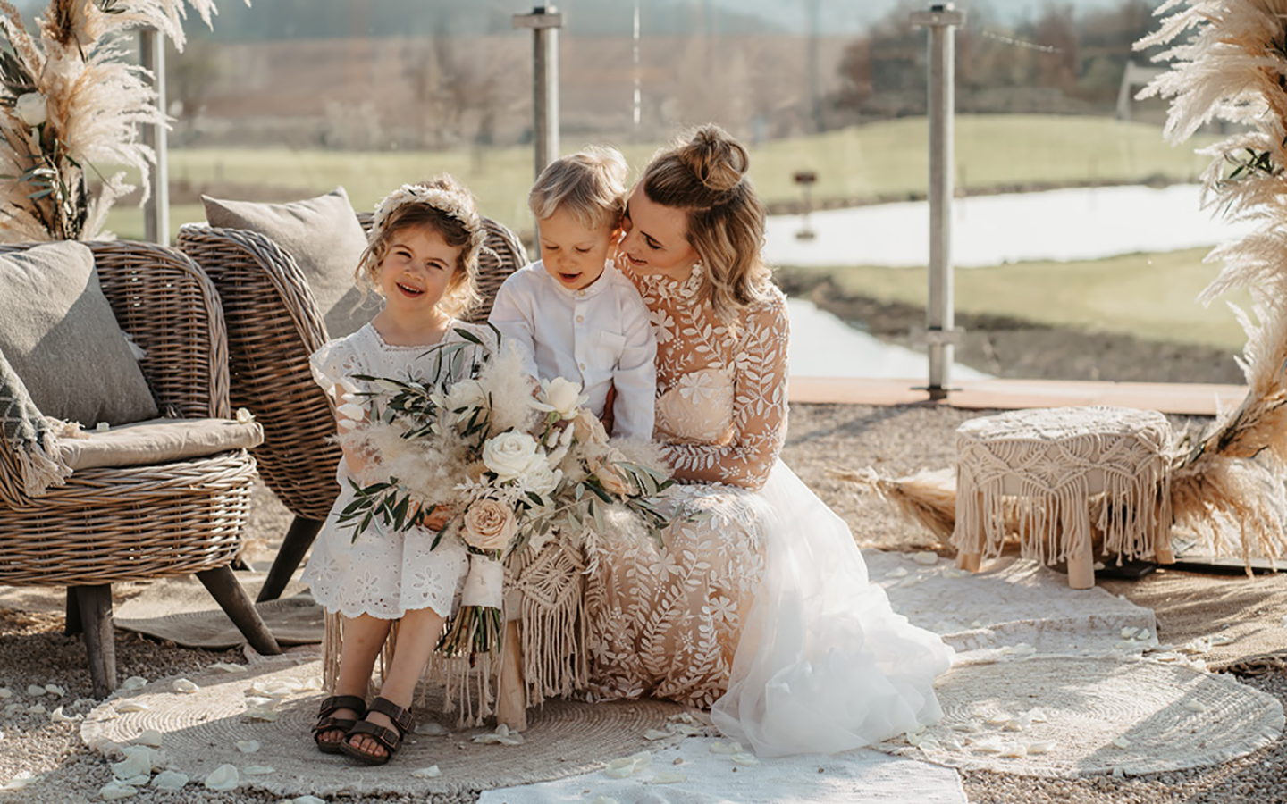 Die Braut und ihre zwei kleinen Kinder sitzen im Freien in der Sonne unterhalb des Traubogens, bereit für Fotos.
