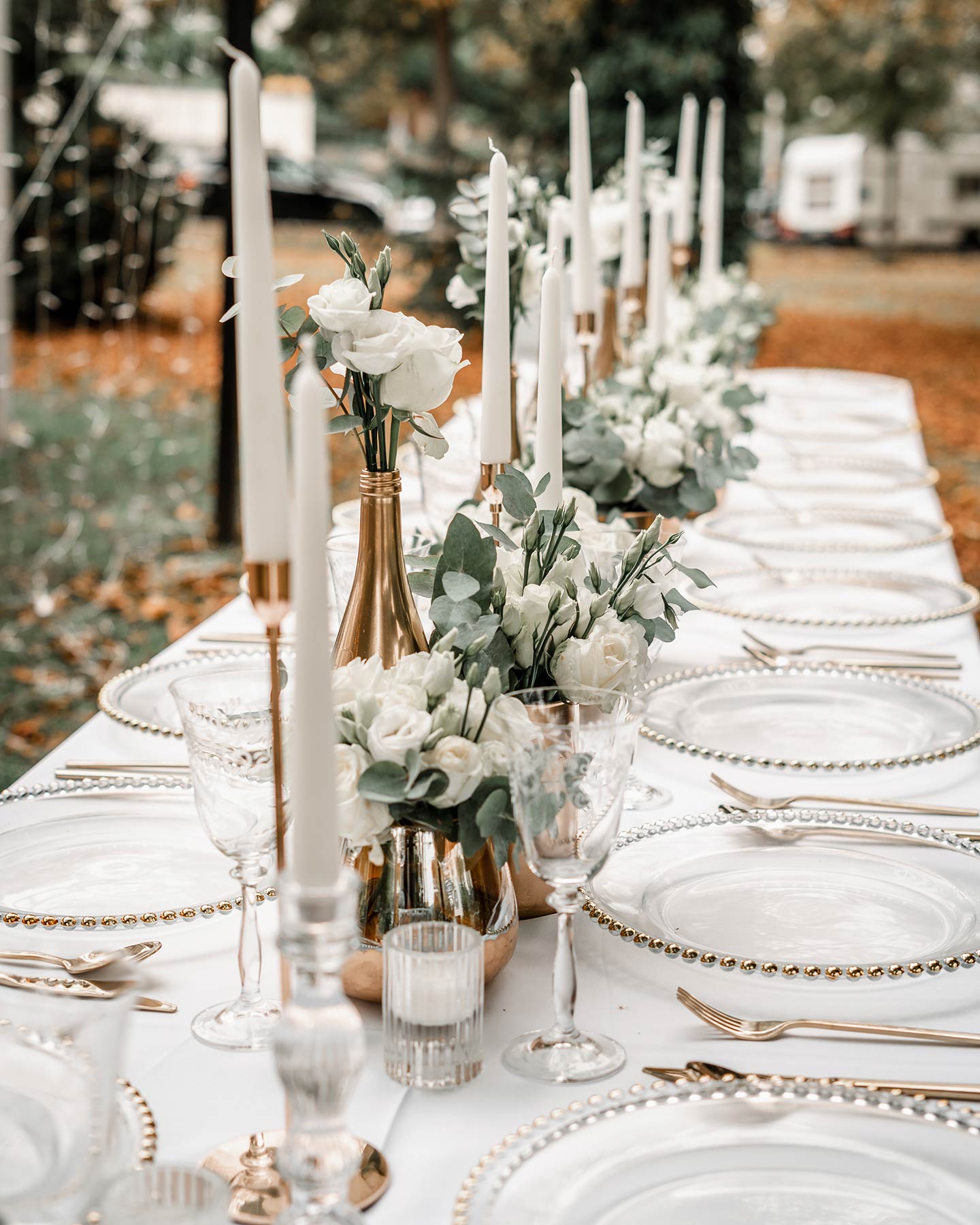 Der gedeckte Hochzeitstisch ist liebevoll angeordnet mit durchsichtigen Tellern mit Goldrand, goldenem Besteck, weißen, großen Kerzen und grün-weißen Blumengestecken.