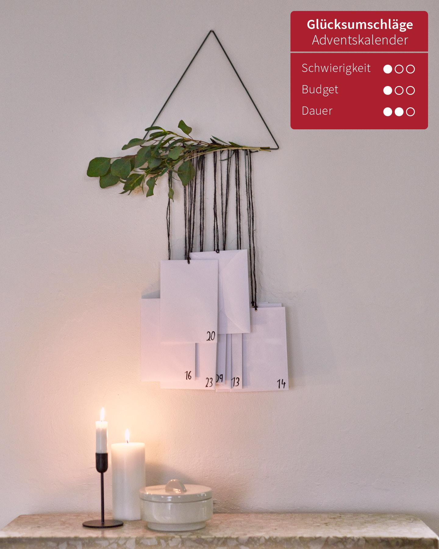 Selbstgemachter Adventskalender mit Umschlägen, die an einem Metalldreieck befestigt sind, hängt als Dekoelement an einer weißen Wand.