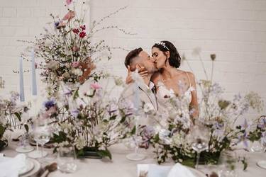 Verliebtes Brautpaar sitzt an Hochzeitstafel, die dekoriert ist mit violetten, weißen und rosa Blumen.