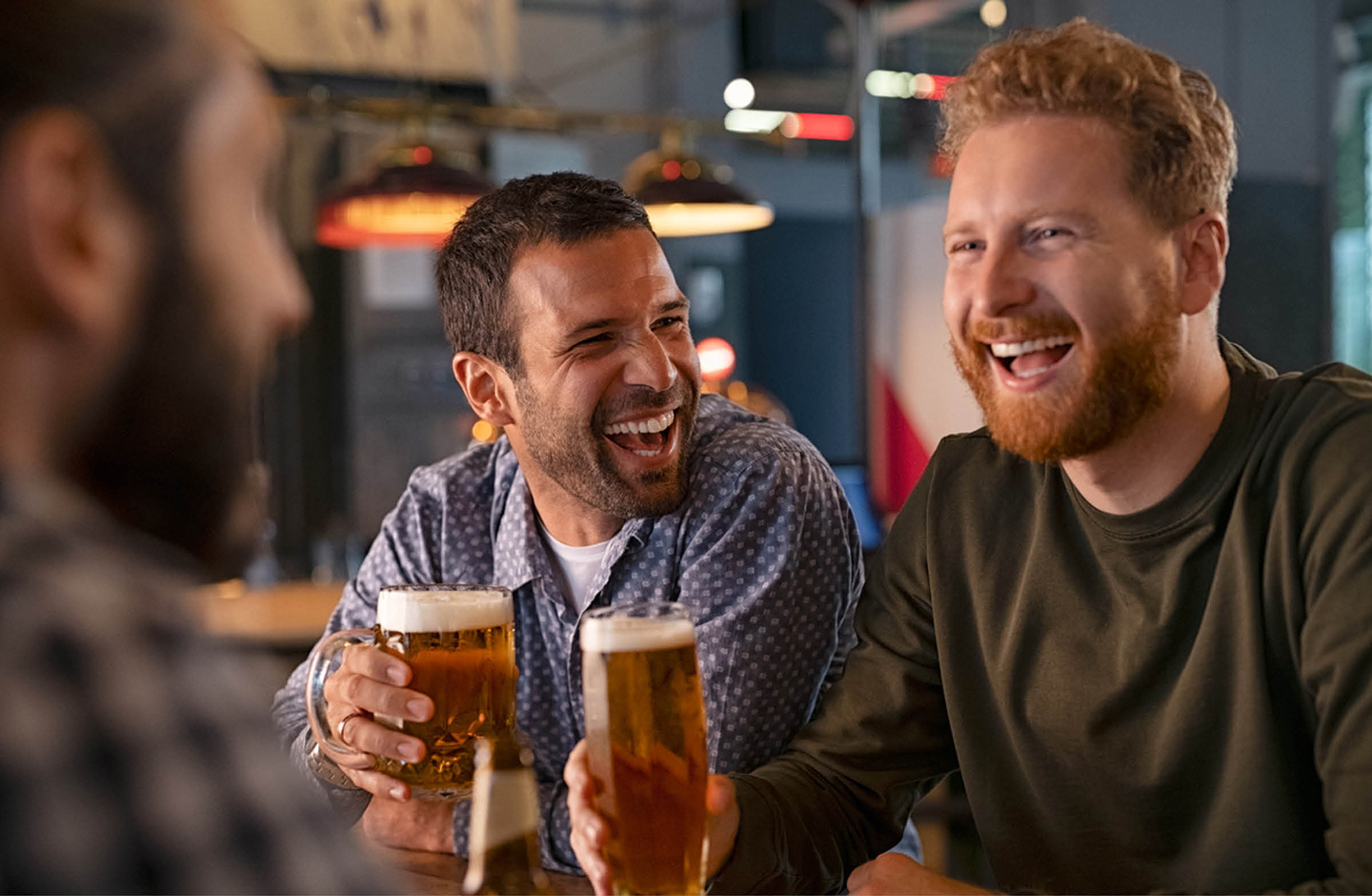 Drei Männer sitzen am Tisch und stoßen mit einem Bier an. Die Stimmung ist heiter.