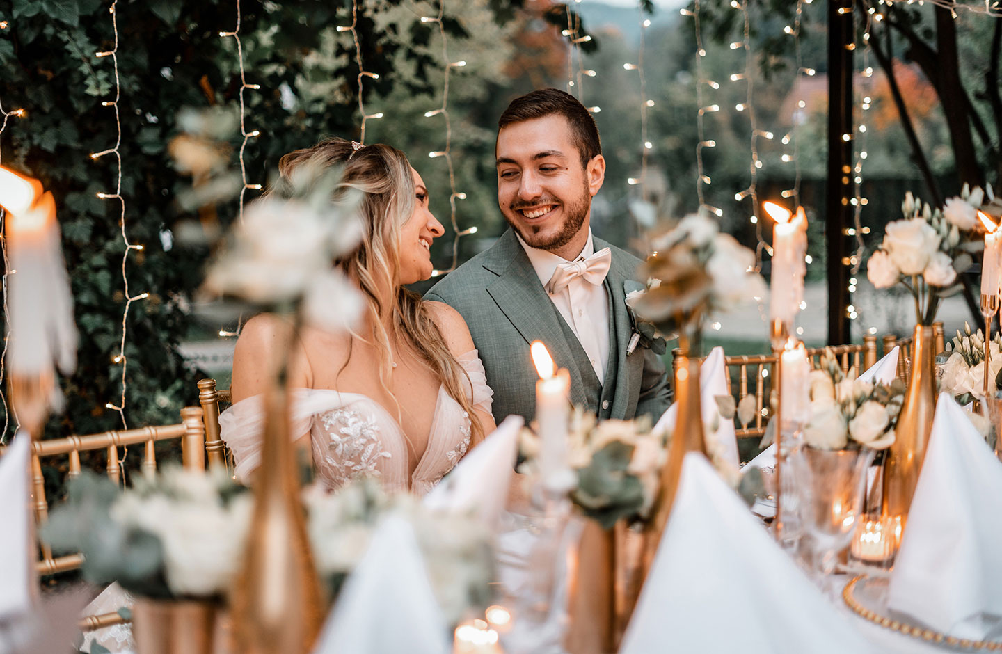 Das Hochzeitspaar aus einer Braut mit einem cremefarbenen, verspielten Kleid und ihrem Bräutigam mit einem grauen Anzug mit cremefarbener Fliege, schaut sich verliebt an. Die beiden sitzen an der liebevoll hergerichteten Hochzeitstafel.