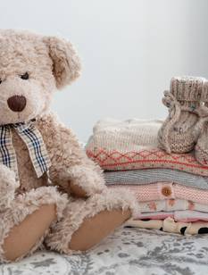 Teddybär neben einem Stapel niedlicher Babykleidung auf einem Bett