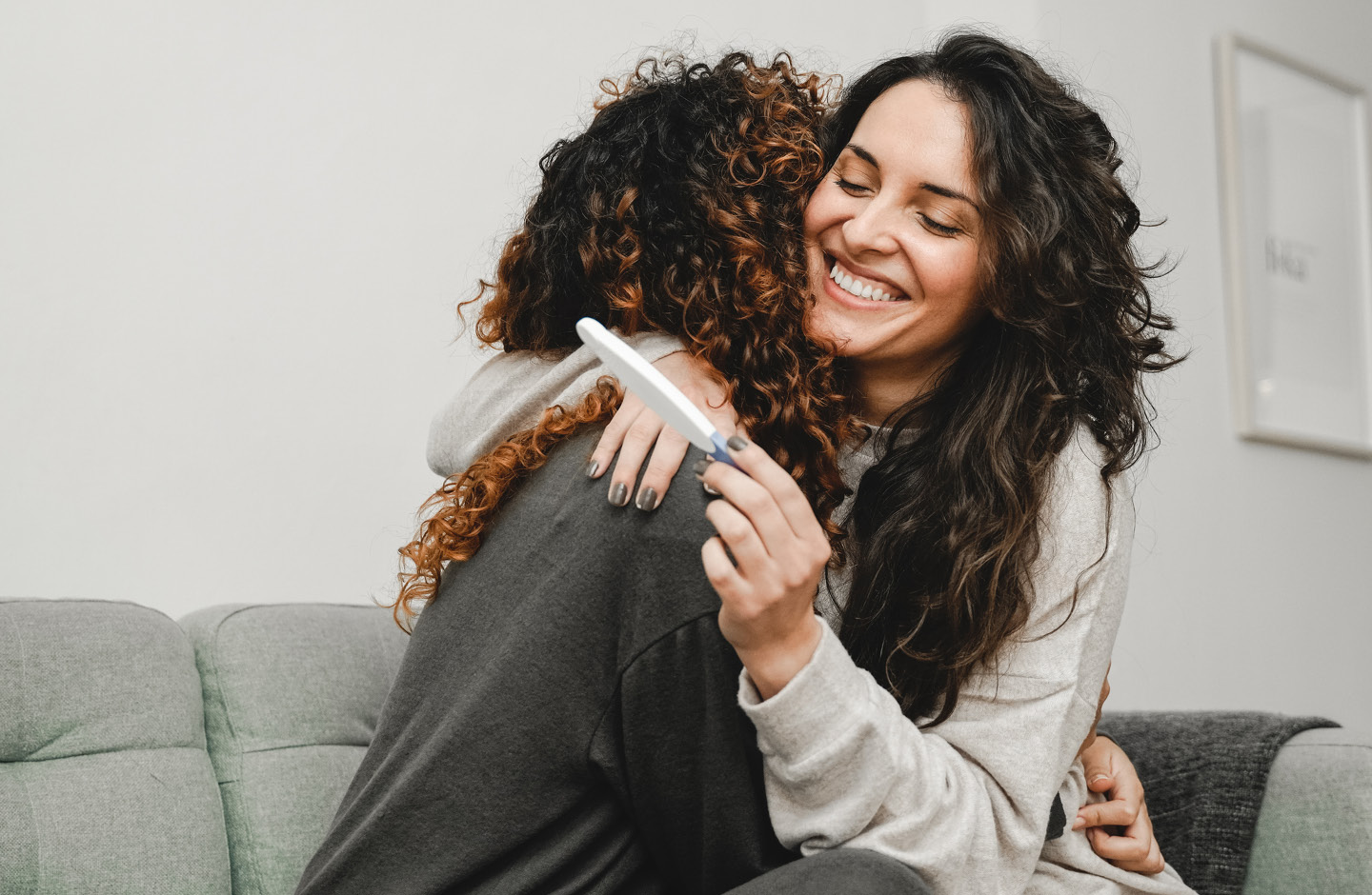 Frau mit positivem Schwangerschaftstest umarmt Freundin auf Sofa