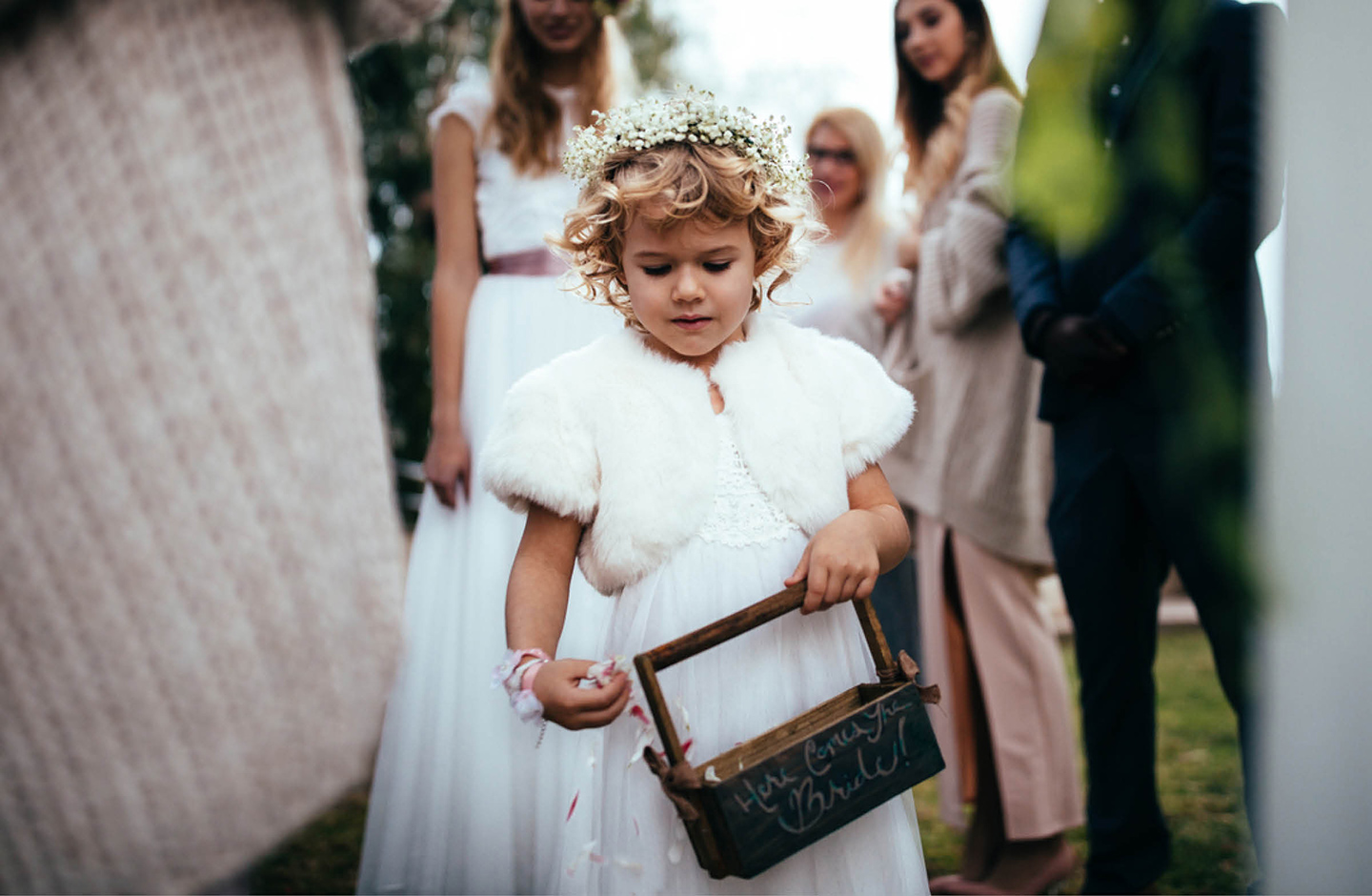 Kleines Mädchen streut Blumen auf einer Hochzeit. Sie trägt ein weißes Kleid und einen Blumenkranz auf dem Kopf. Im Hintergrund sind die Braut und die Hochzeitsgäste in Umrissen zu sehen.