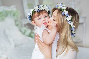 Mama mit Blumenkranz im Haar küsst ihr Baby am Tag der Taufe liebevoll auf die Wange.