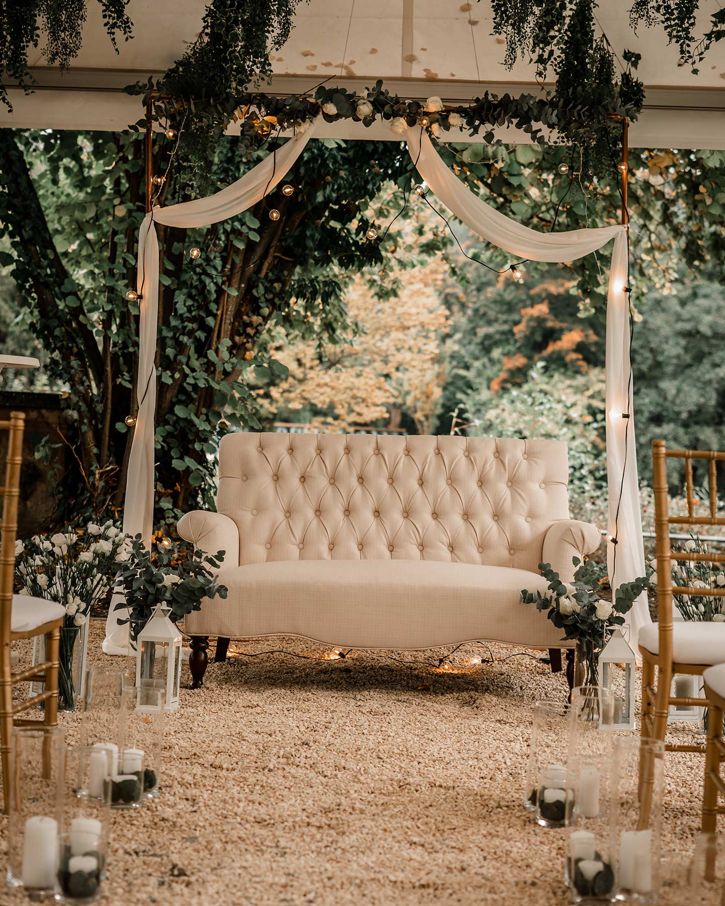 Unter einem weißen Pavillon überdacht steht ein cremefarbenes Sofa für das Hochzeitspaar zur Trauung bereit. Der Traubogen in Gold wird von einer Girlande aus weißem, dünnen Stoff, einer Lichterkette und einer grünen Pflanzenranke verziert.
