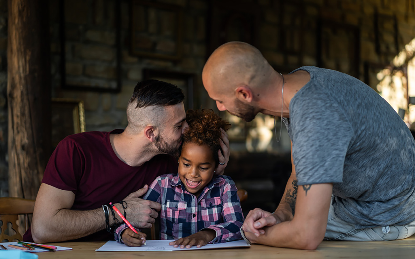 Zwei Väter sitzen mit ihrer Tochter am Tisch und unterstützen sie bei ihren Hausaufgaben.
