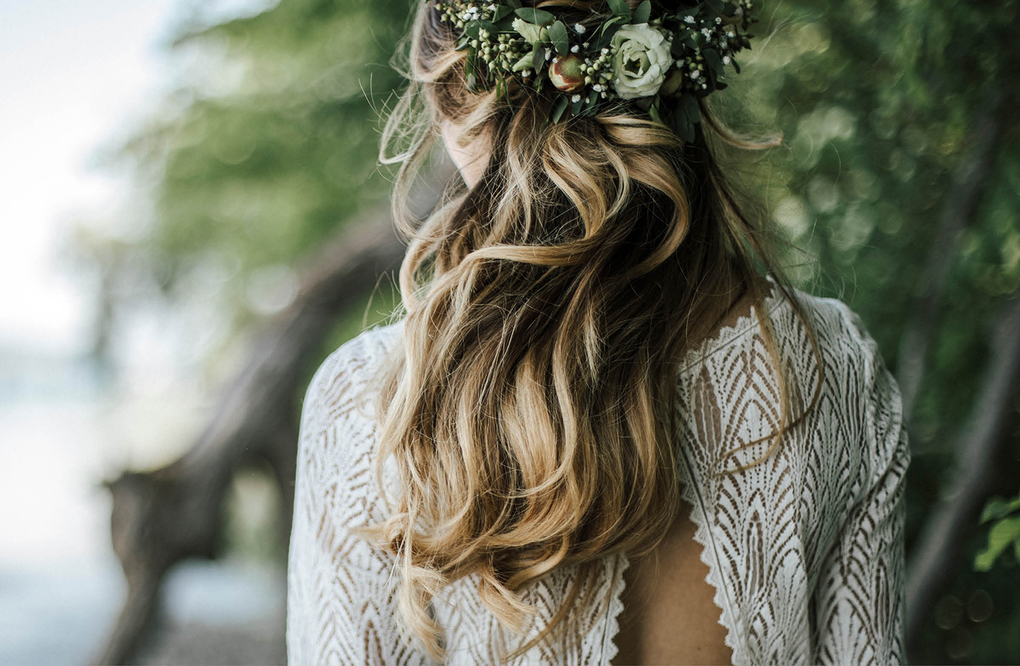 Rückenansicht der Braut. Die Braut trägt die Haare offen und leicht gewellt mit einem Blumenkranz. Das Hochzeitskleid hat einen Rückenausschnitt