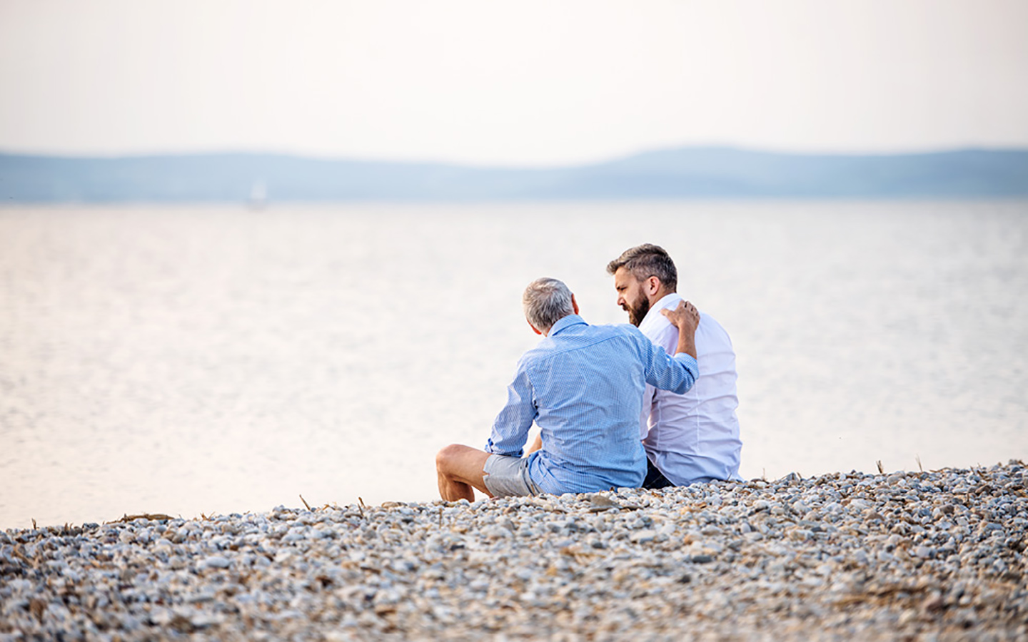 Vater und Sohn sitzen am Meer und schauen gemeinsam auf das Wasser. Der Vater hält schützend und gut zuredend seine Hand auf den Rücken seines Sohnes.