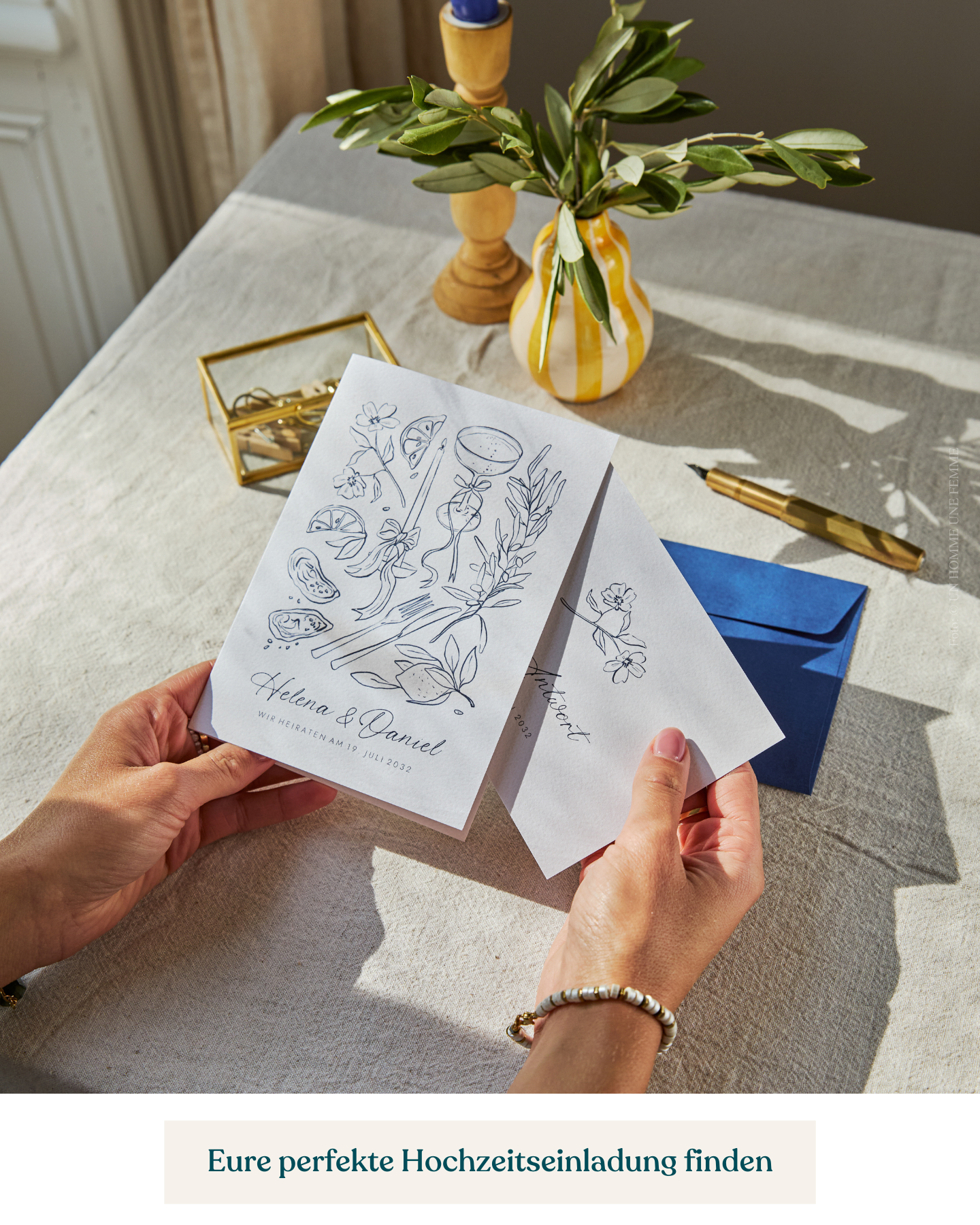 Frau (nicht im Bild9 hält Hochzeitseinladung mit Antwortkarte, sie sitzt an einem Tisch mit beigefarbener Leinendecke und mediterraner Deko