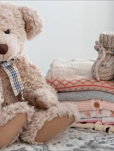 Teddybär neben einem Stapel niedlicher Babykleidung auf einem Bett