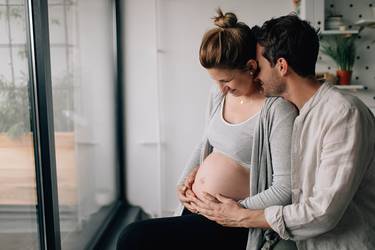 Mann umarmt seine schwangere Frau von hinten und hält ihren Babybauch. Er legt seinen Kopf auf die Schulter seiner Frau.