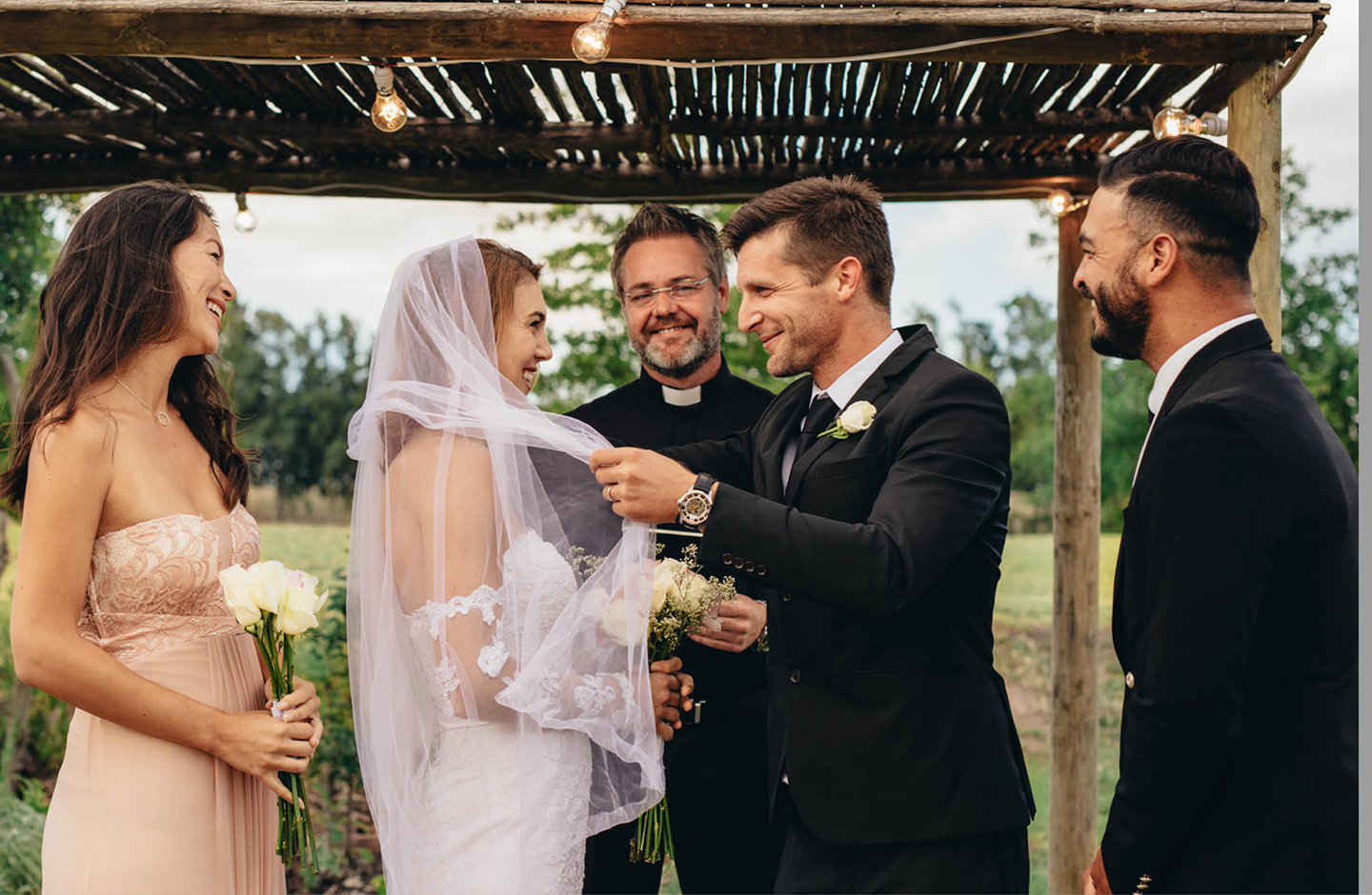 Bräutigam nimmt Braut den Schleier ab. Der Trauredner steht im Hintergrund. Links und rechts neben dem Brautpaar stehen die Trauzeugin und der Trauzeuge.