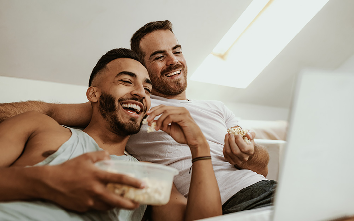 Ein glückliches Paar bestehend aus zwei Männern liegt auf dem Sofa und schaut erfreut, mit Popcorn in der Hand, auf den Laptop-Display. 