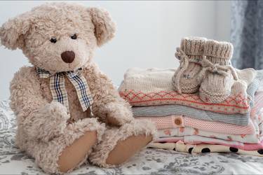 Teddybär neben einem Stapel niedlicher Babykleidung auf einem Bett