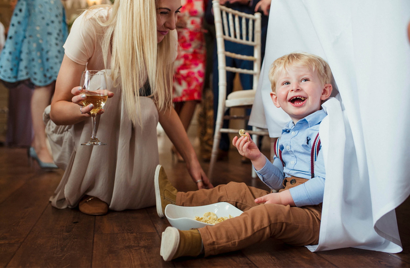 Ein kleiner Junge sitzt auf dem Boden der Hochzeitslocation unter einem Tisch. Er lacht und nascht Süßigkeiten aus einer Schüssel. 