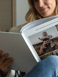 Junge Frau blättert lächelnd in einem Fotobuch mit Hardcovereinband, das Cover zeigt eine Collage mit Familienfotos