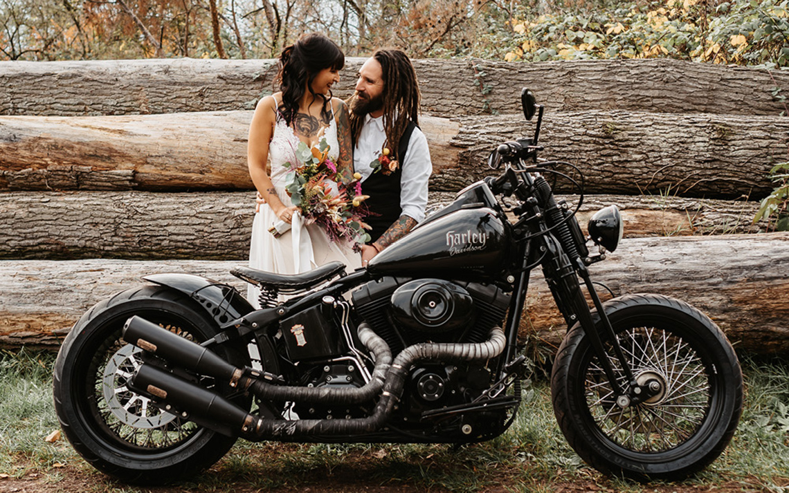 Frisch getrautes Pärchen vor Kulisse aus Baumstämmen in der Natur, im Hochzeitsoutfir mit Brautstrauß und Harley im Vordergrund.