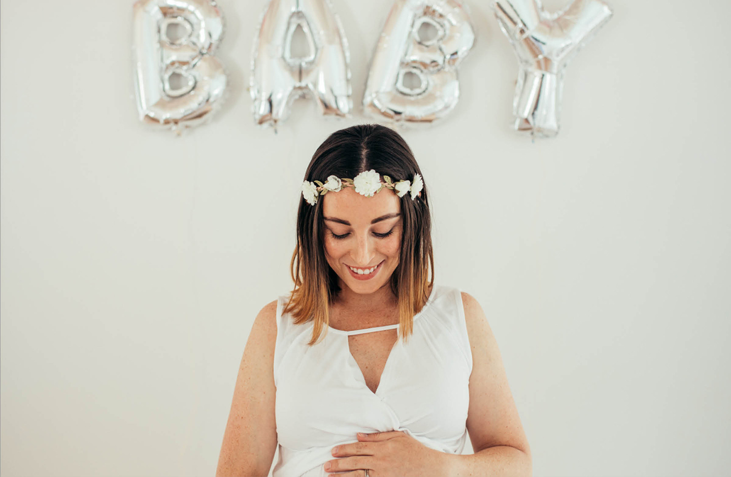 Schwangere Frau mit Blumenkranz im Haar hält ihren Baby Bauch. An der Wand im Hintergrund sind silberne Folienballons mit dem Schriftzug Baby befestigt. 