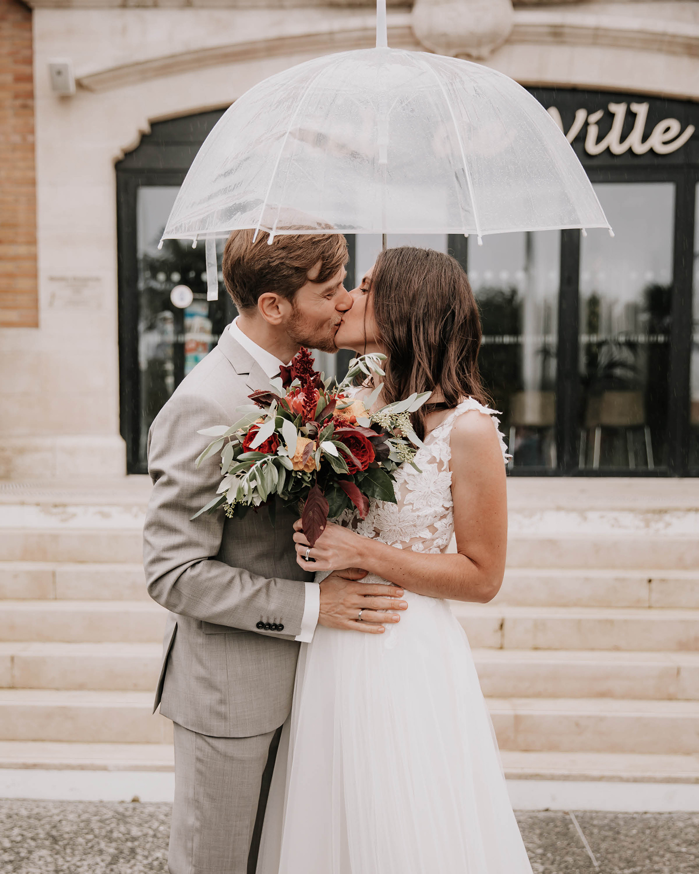 Ein sich küssendes Hochzeitspaar steht vor der Location. Über ihren Köpfen halten sie einen Regenschirm.