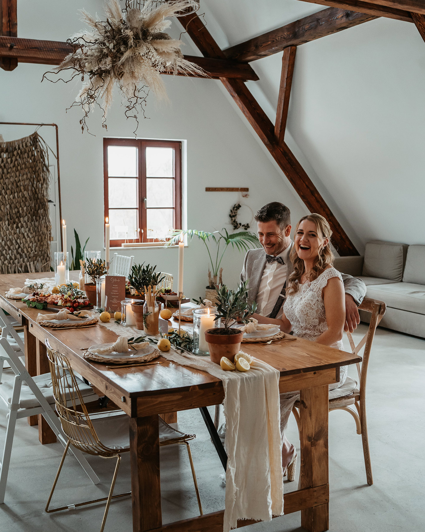 Braut und Bräutigam sitzen an ihren Plätzen an der Hochzeitstafel. Die rustikale Tafel ist mit Greenery-Elementen wie Eukalyptus geschmückt. Windlichter sorgen für romantische Stimmung.