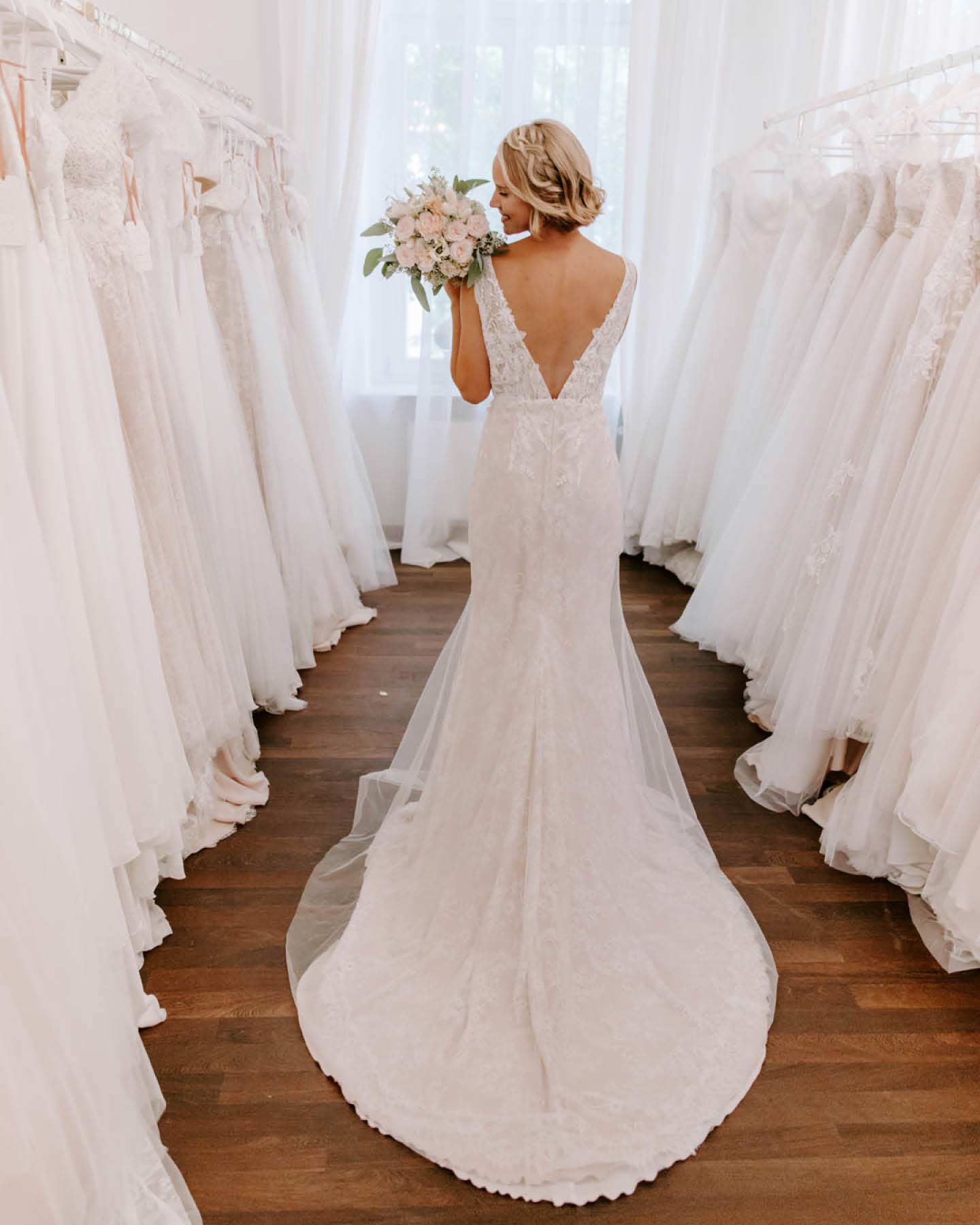 Braut steht im Brautmodenladen und zeigt den tiefen Rückausschnitt ihres Brautkleides.