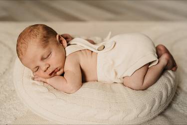 Niedliches Baby in Latzshorts schläft bäuchlings auf einem cremefarbenen Kissen Kissen