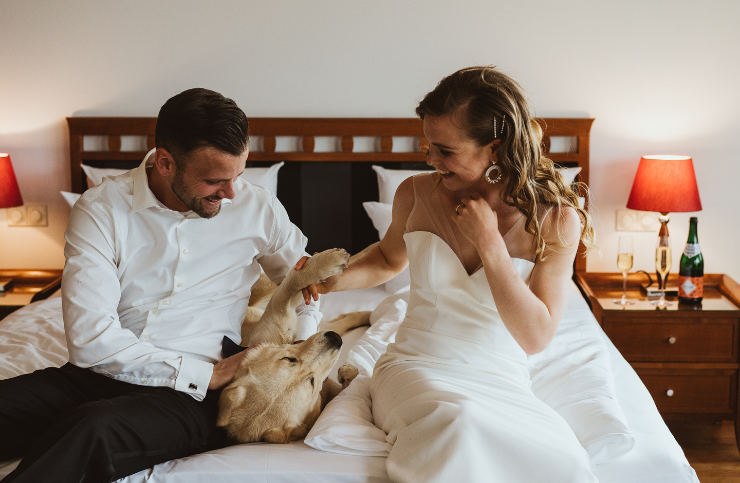 Angehendes Ehepaar sitzt im Hochzeitsoutfit auf dem Bett und spielt mit dem Hund
