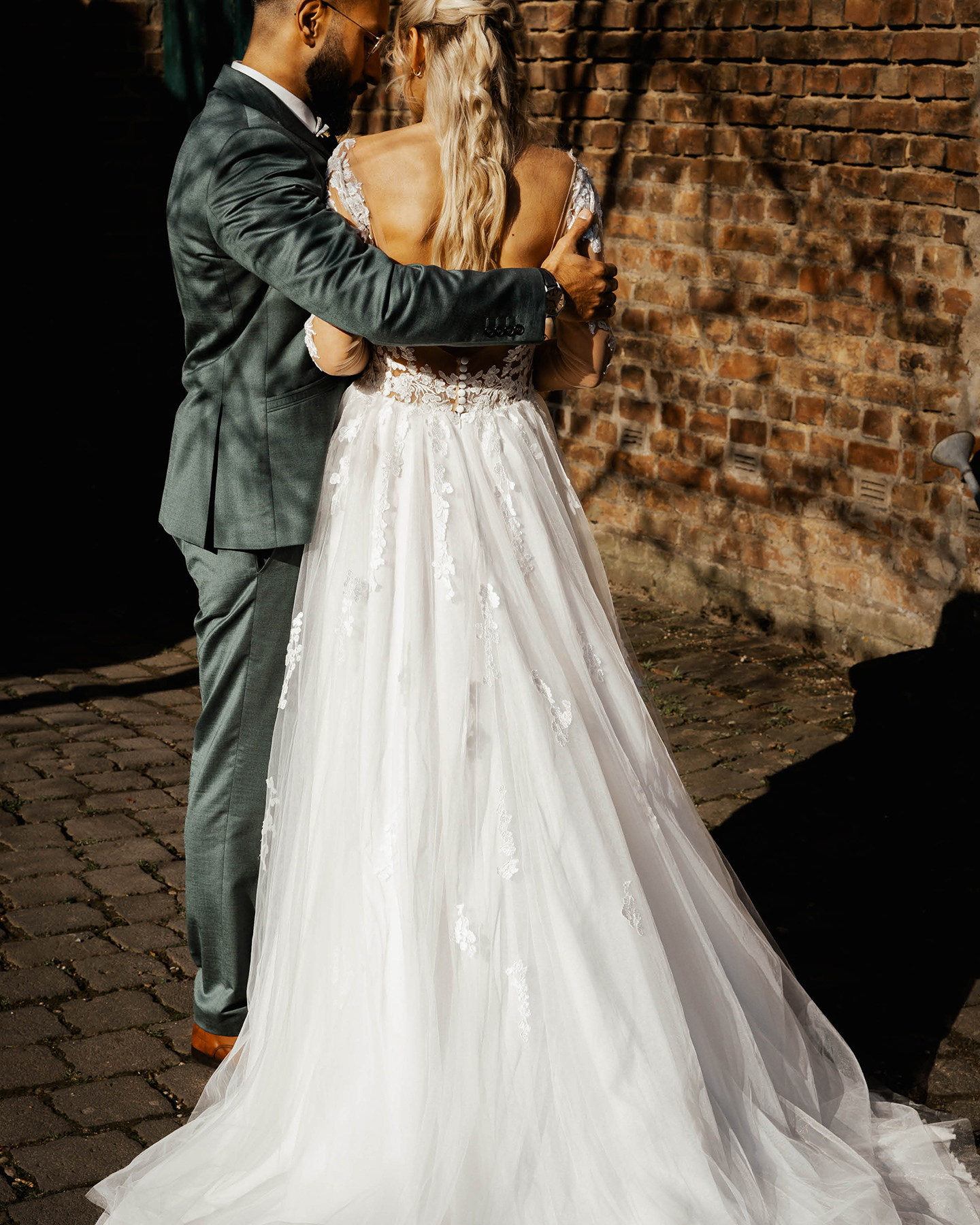  Bräutigam im blaugrauen Anzug legt Arm um seine Braut, die ein Kleid aus weißer Spitze und Chiffon trägt. Im Hintergrund eine Backsteinmauer.
