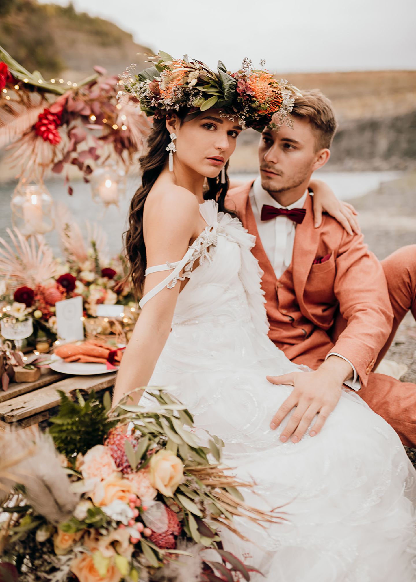 Das Brautpaar sitzt auf der selbstgemachten Hochzeitstafel, die in Rot- und Orangetönen geschmückt ist. Die Braut hat eine Blumenkrone auf ihrem Kopf.