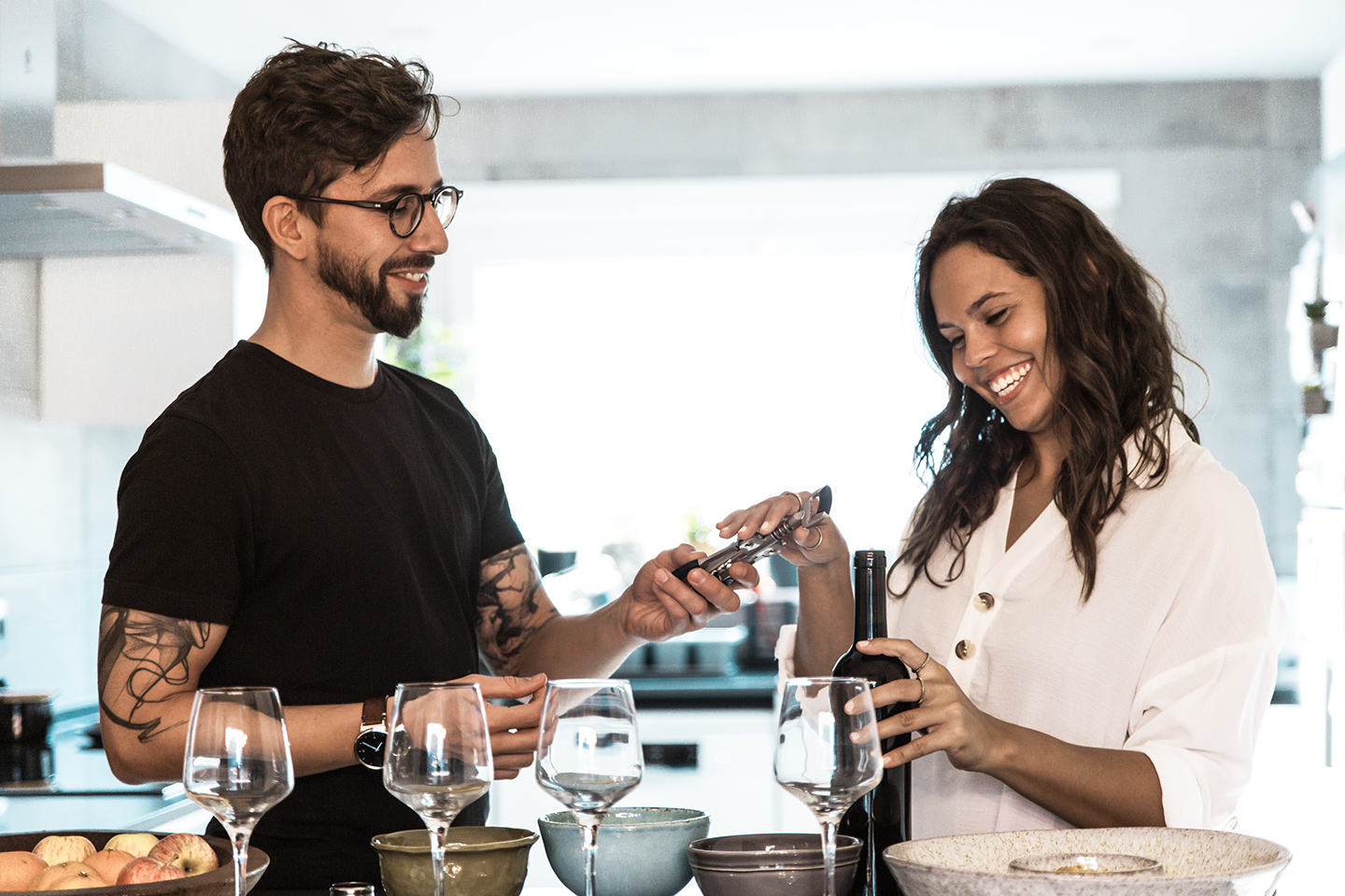 Ein Tasting für Wein findet bei diesem Paar in der eigenen Küche statt. Mann und Frau freuen sich über die Weinflasche und die Gläser vor sich in der Küche und sind voller Vorfreude auf diese Geschenkidee.