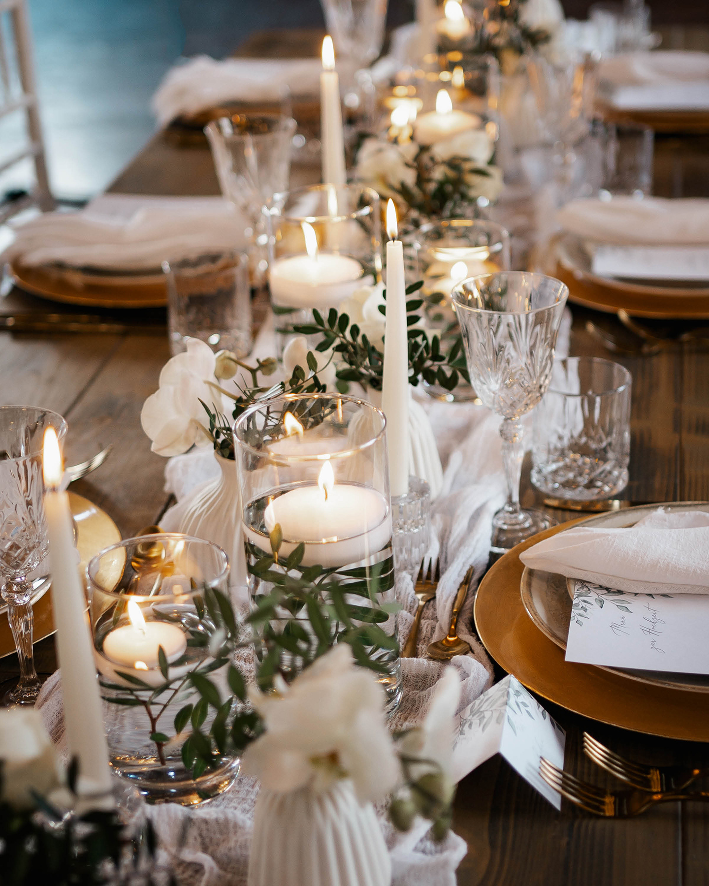 Festlich gedeckte Hochzeitstafel mit weißen Kerzen, hellen Stoffen und weißen und grünen foralen Akzenten.