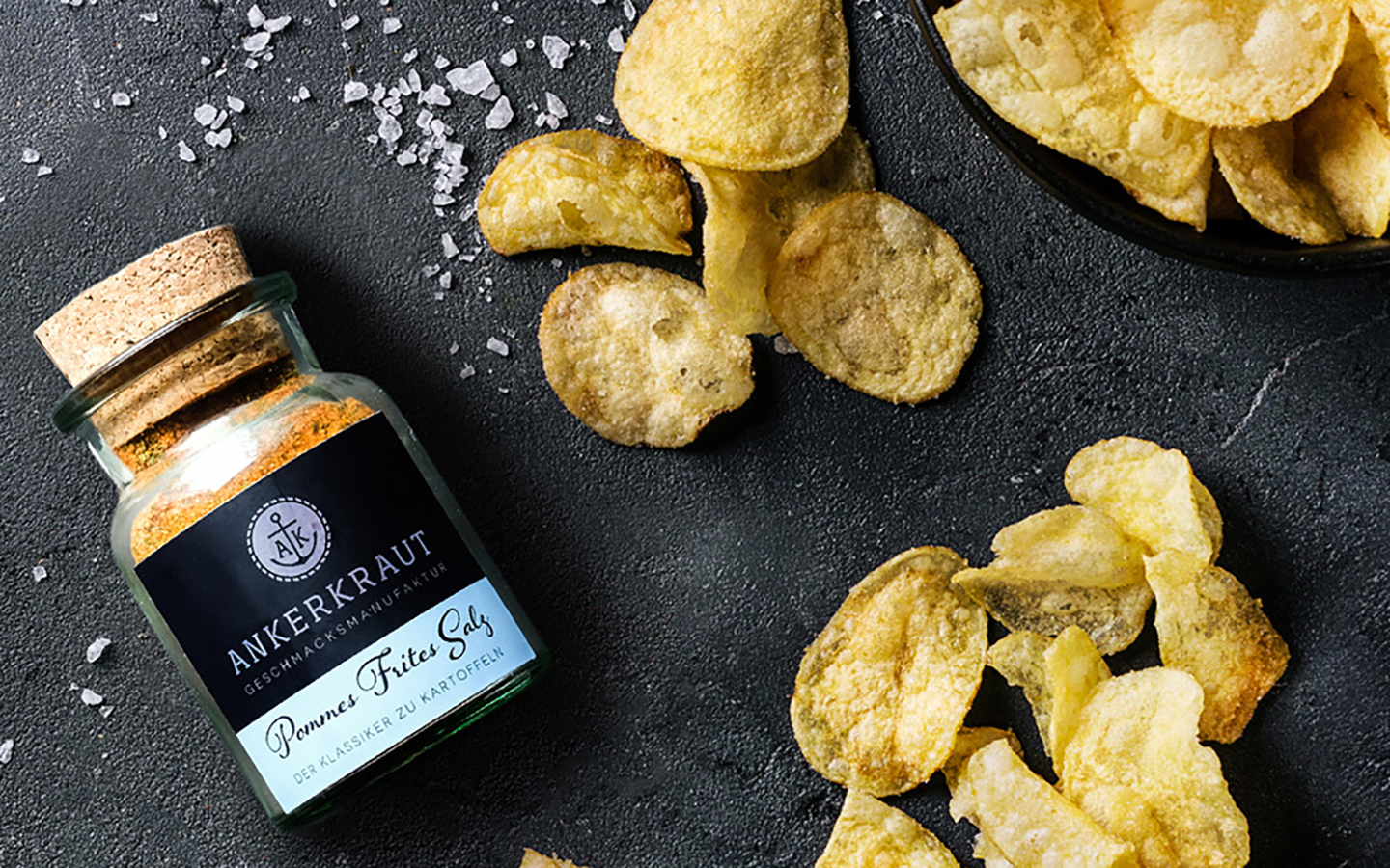 Gewürzmischung "Pommes Frites Salz" von Ankerkraut liegt auf einer schwarzen Unterlage neben den fertig selbstgemachten Kartoffelchips aus dem Ofen.
