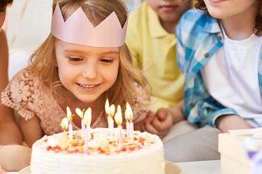 Geburtstagskind mit Krone auf auf dem Kopf sitzt mit seinen Geburtstagsgästen am Tisch und pustet die Kerzen auf der Torte aus