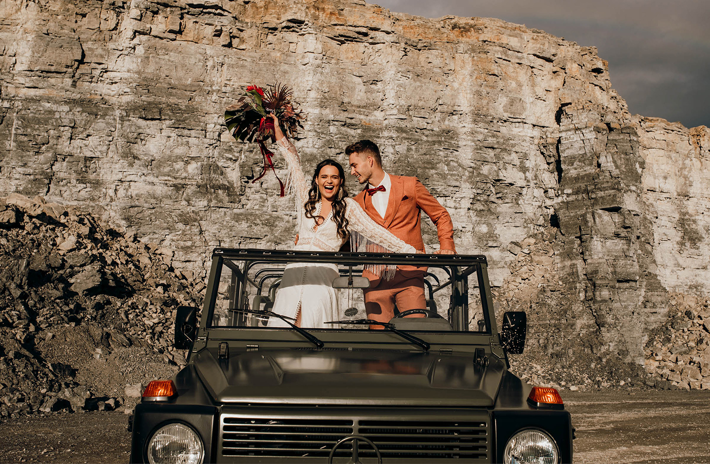 Brautpaar steht in einem Geländewagen. Die Braut hält ihren Brautstrauß in die Luft. Im Hintergrund sieht man die Steinmauern eines Steingutes 